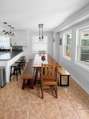 Une cuisine avec des meubles blancs, une table à manger en bois et un banc. On y trouve des luminaires suspendus et une fenêtre avec des stores.