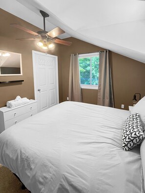 Un dormitorio con cama, ventilador de techo, ventana con cortinas, mesita de noche y espejo.