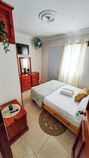 Quarto com cama, cômoda, espelho, televisão e ventilador de teto.