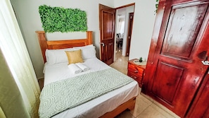 A bedroom with a bed, a wooden headboard, a green wall decoration, a wooden door, and a nightstand with a plant.