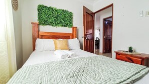 A hotel room with a bed, a wooden headboard, a green wall decoration, a bedside table with a plant, and a door leading to a bathroom.