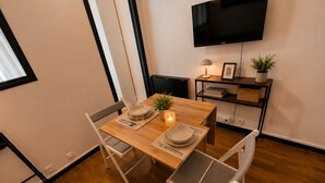 A dining area with a wooden table, white chairs, a TV mounted on the wall, and a small plant on a shelf. - Cozy apartment just outside Paris – Clichy (Clichy)