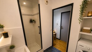A compact bathroom with a glass shower enclosure, a sink, a toilet, and a washing machine. - Cozy apartment just outside Paris – Clichy (Clichy)