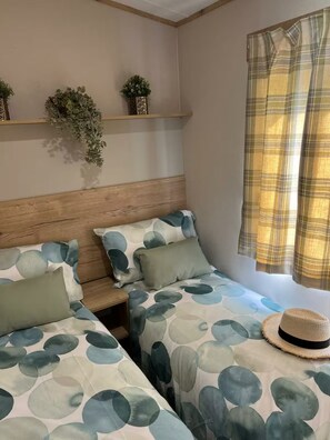 Ein Zimmer mit zwei Betten, einem hölzernen Kopfteil, einem Regal mit Pflanzen und einem Fenster mit einem Karomuster-Vorhang.