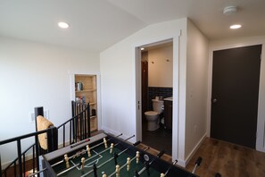 Una habitación con mesa de fútbol, estantería y un baño visible a través de una puerta abierta.