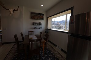 Een eethoek met een tafel, stoelen, een opgehangen hertenschedel, een raam met uitzicht en een koelkast.