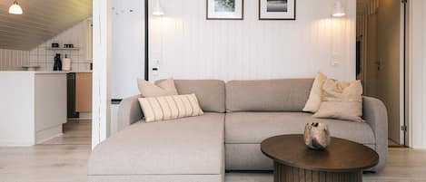 Una sala moderna con un sofá modular, una mesa de centro redonda y una cocina al fondo.