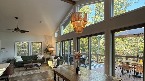 Amplia sala de estar con una gran mesa de madera, un sofá y un comedor con sillas. Amplias ventanas ofrecen vistas al exterior.