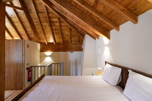 Une chambre avec un plafond en bois, un lit recouvert de draps blancs, une table de chevet avec une lampe et un escalier menant à une autre pièce.