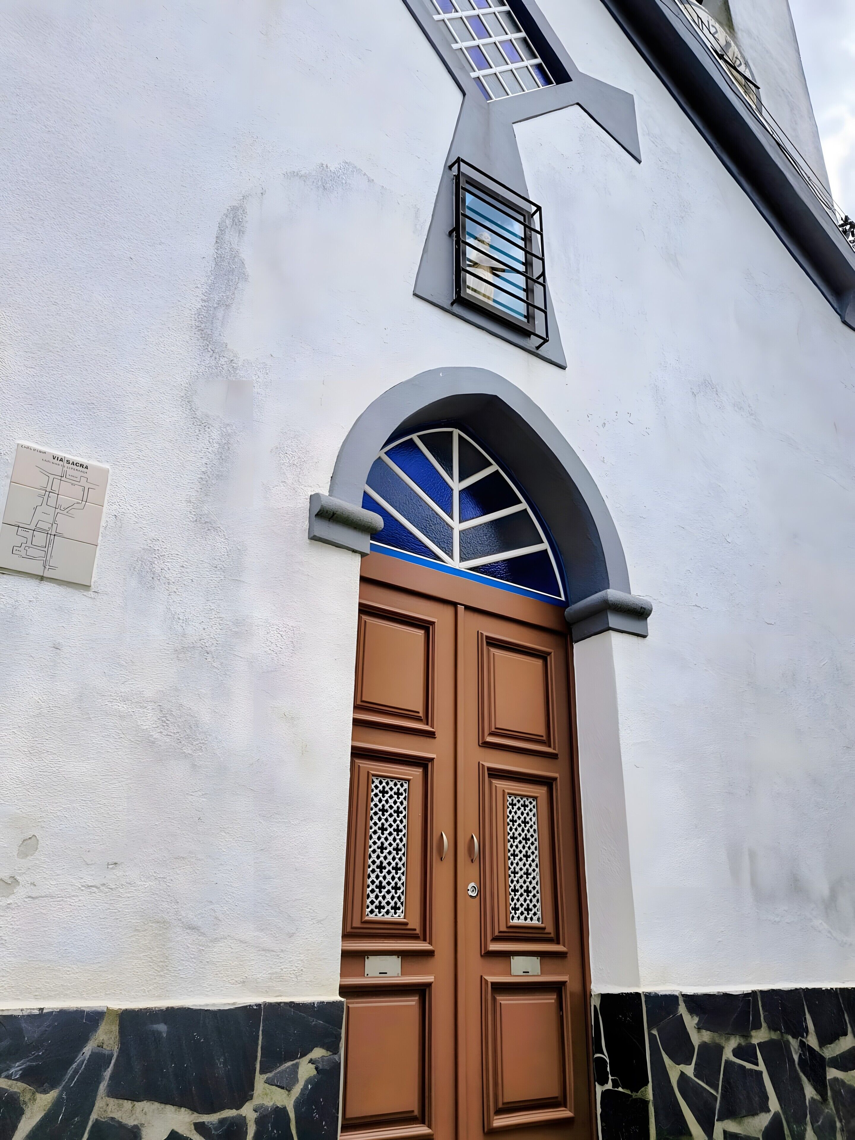 Un bâtiment à la façade blanche, doté d’une porte à double battant de couleur marron ornée de panneaux décoratifs, et d’une fenêtre aux carreaux bleus située au-dessus de la porte.