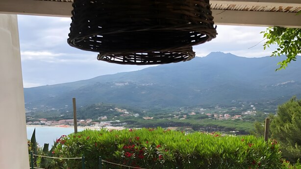 A patio with a table and chairs, a wicker chair, and a view of a mountain and beach. - Holiday Home 'Terrazza sul Mare' with Sea View and Private Terrace (Marina di Campo)