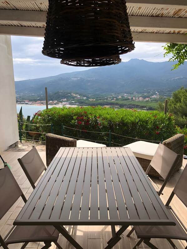 Un patio con una mesa y sillas, una silla de mimbre, y vistas a la montaña y la playa.