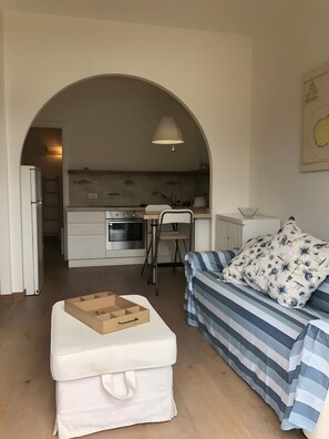 A compact living space with a kitchenette, a white sofa with patterned cushions, and a wooden ottoman. - Holiday Home 'Terrazza sul Mare' with Sea View and Private Terrace (Marina di Campo)