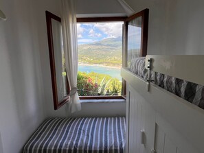 A room with a window view of a coastal landscape, a bed with striped bedding, and a white wall with a textured panel. - Holiday Home 'Terrazza sul Mare' with Sea View and Private Terrace (Marina di Campo)