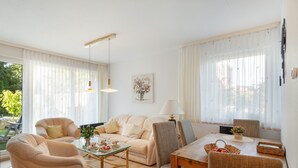 Een goed verlichte woonkamer met een bank, fauteuils, een glazen salontafel en een eettafel met rieten stoelen.