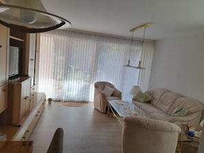 Una sala de estar con un sofá, una mesa de centro, un televisor y un ventanal con cortinas de gasa.