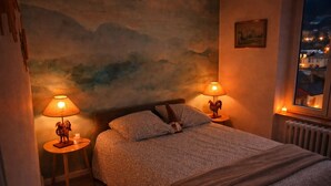A bedroom with a bed, two bedside lamps, a wall mural, a window with a view, and a radiator. - Mountain Charm in La Bourboule - Apartment with Terrace and Small Garden (La Bourboule)