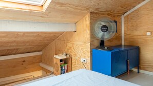 Ein gemütlicher Dachraum mit schrägen Wänden, einem kleinen Holzregal mit Büchern, einem blauen Schrank und einem Standventilator.