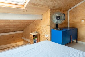 A compact attic room with a sloped ceiling, a small wooden shelf with books, a blue cabinet, and a standing fan. - Stay in the Heart of Capbreton with Private Terrace (Capbreton)