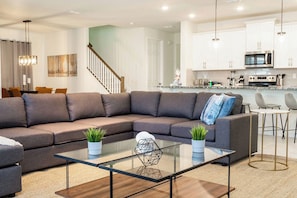 A modern living room with a sectional sofa, a glass coffee table, and a kitchen area with white cabinets and stainless steel appliances. - Modern 9BR Family Villa Pool and Relax (Kissimmee)