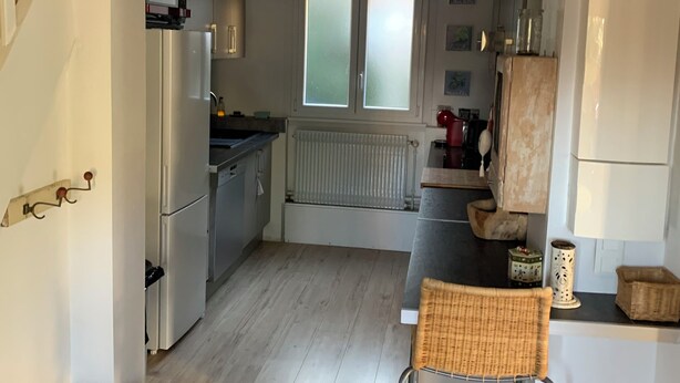 Eine Küche mit Kühlschrank, ein Essbereich mit einer Bank und Stühlen, eine Treppe und ein Fenster mit Milchglas.