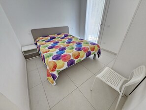 Ein Bett mit farbig gepunktetem Bettbezug, ein weißer Stuhl und ein kleiner Tisch in einem Zimmer mit weißen Wänden und einem gefliesten Boden.