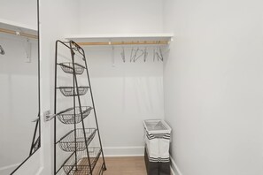 A white room with a metal rack holding baskets, a wooden shelf, and a small table with folded white cloths. - Kali House •  River Ranch • Prime Location • Premier Host (Lafayette)