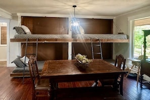 A room with a central wooden dining table, two bunk beds with wooden frames, and a hanging light fixture.