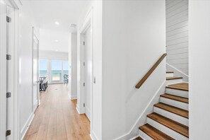 Un couloir menant à un balcon d’où l’on peut admirer la plage, un escalier en bois et des murs blancs.