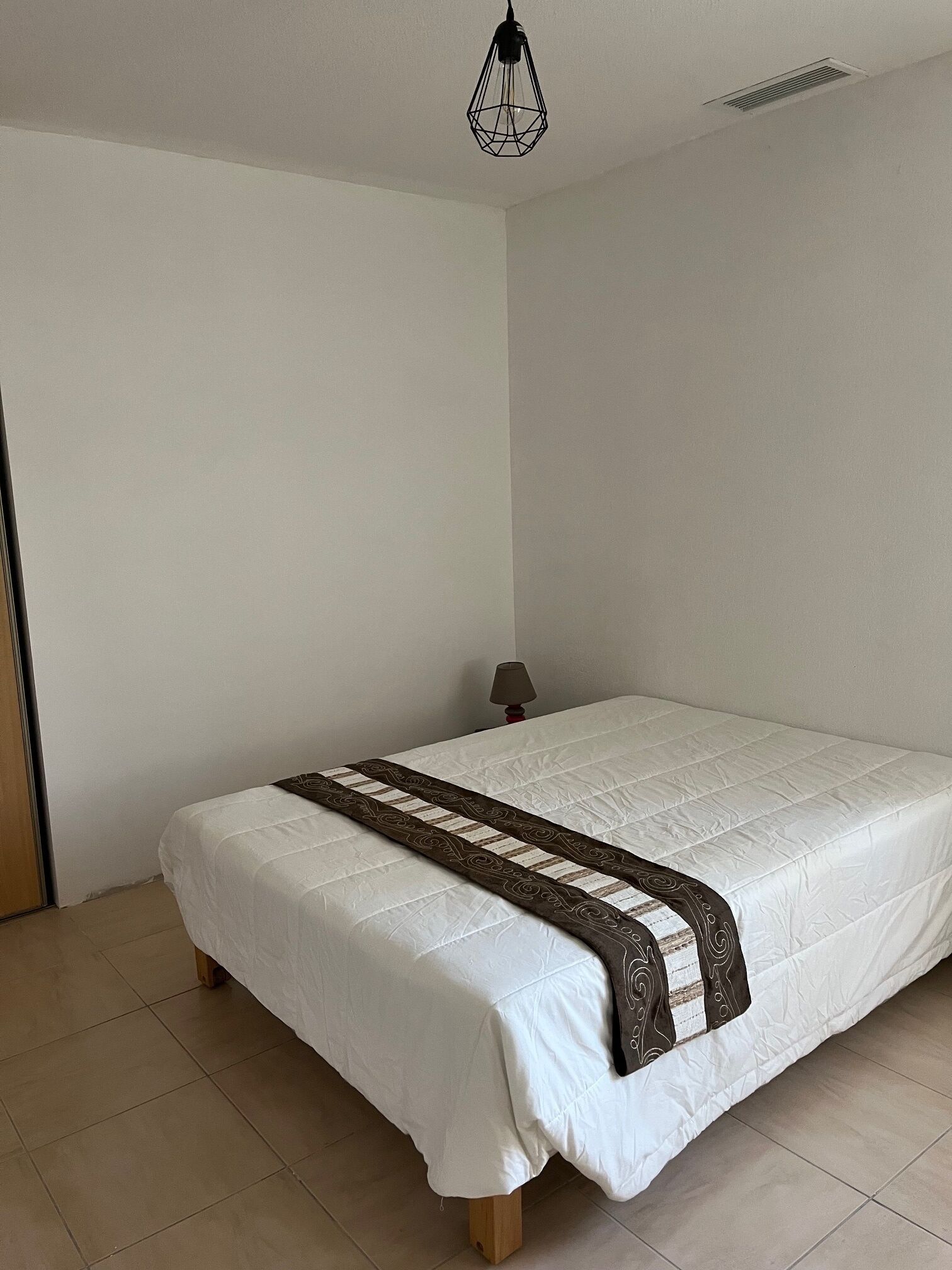 Uma cama de solteiro com colchão branco e manta decorativa marrom, luminária de cabeceira e luminária de teto geométrica.
