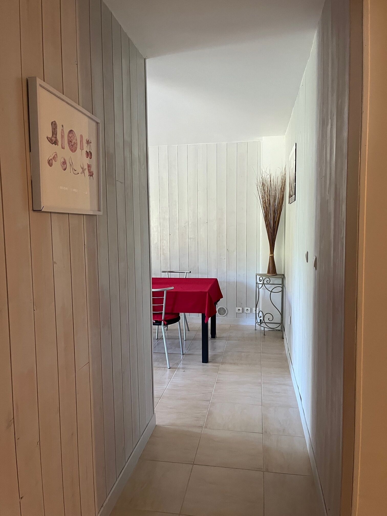 Een gang met een eethoek, met een rode tafel en stoelen, een ingelijst beeld aan de muur, en een vaas met hoge planten.