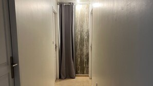 Un couloir étroit au sol carrelé, avec une porte sur la gauche et une fenêtre recouverte d’un rideau sur la droite.