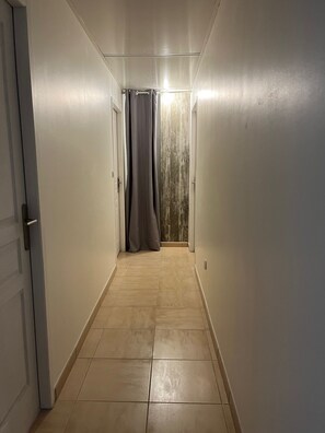 Un couloir étroit au sol carrelé, avec une porte sur la gauche et une fenêtre recouverte d’un rideau sur la droite.