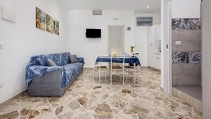 Una habitación con piso de piedra, un sofá, una mesa de comedor con sillas, un televisor de pantalla plana y una pequeña cocina.