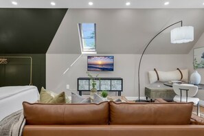 Ein modernes Wohnzimmer mit einem braunen Ledersofa, einer Glasvitrine, einem Sonnenuntergangsbild, einer Stehlampe und einem Fenster mit Blick ins Freie.