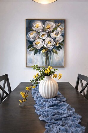 A dining table with a white vase and yellow flowers, a blue ruffled table runner, and a painting of white flowers on the wall. - Near Niagara Falls+Wine Country+Games Tables (Thorold)