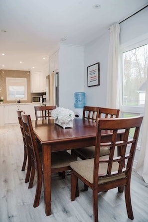 A dining area with a wooden table and chairs, a water dispenser, and a kitchen in the background. - Wylde Roots Escape - Hidden Beachside Cottage (Port Colborne)