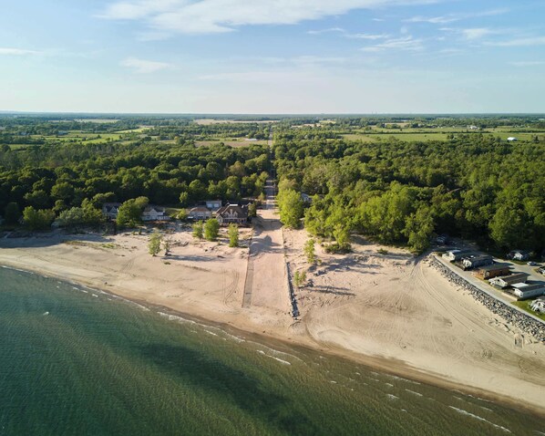 An aerial view of a coastal area with a beach, a cluster of houses, and surrounding forest. - Wylde Roots Escape - Hidden Beachside Cottage (Port Colborne)