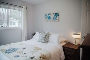 A bedroom with a bed, a nightstand, a lamp, a window, and a wall art piece. - Wylde Roots Escape - Hidden Beachside Cottage (Port Colborne)