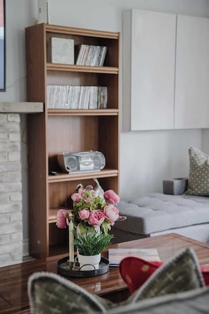A living room with a bookshelf, a radio, a coffee table with a flower arrangement, and a grey sofa. - Wylde Roots Escape - Hidden Beachside Cottage (Port Colborne)