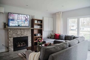 A living room with a fireplace, a large flat-screen TV, a sectional sofa, and a bookshelf. - Wylde Roots Escape - Hidden Beachside Cottage (Port Colborne)