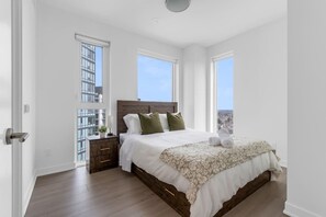 A bedroom with a large bed, two windows, a nightstand, and a view of a cityscape. - 2 BR Apartment at Station Park with Swim Spa (Kitchener)