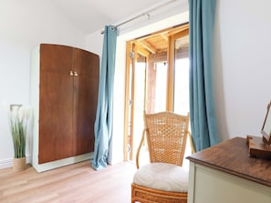 A room with a wooden wardrobe, a wicker chair, a small cabinet, and a view through a door.