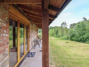 Une terrasse couverte avec des murs en briques et un plafond en bois, des sièges d’extérieur et une vue sur un espace verdoyant avec des arbres.
