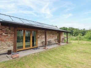 A stone building with large glass doors, a covered patio with chairs, and a grassy yard.