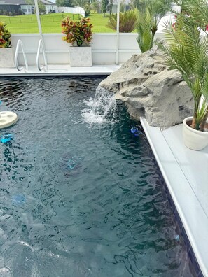 A swimming pool with a waterfall feature, surrounded by potted plants and a view of a green lawn.