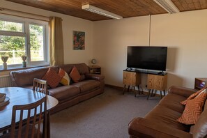 A living room with a brown leather sofa, a wooden dining table, a TV on a stand, and a window with curtains. - Grapnells Lodge, Wallasea Island, Rochford, Essex (Rochford)