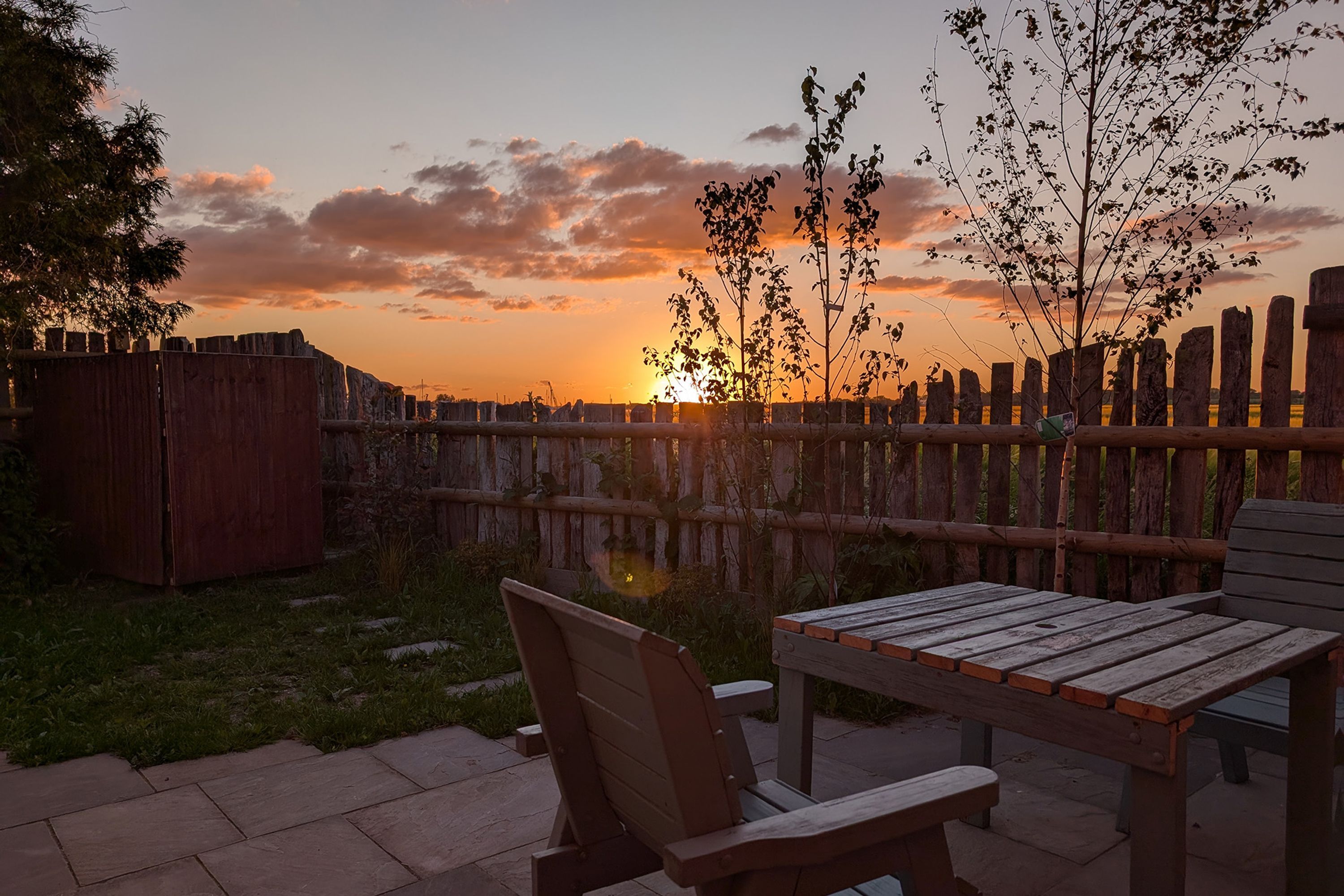 An outdoor seating area with a wooden table and chairs, a wooden fence, and a sunset in the background.