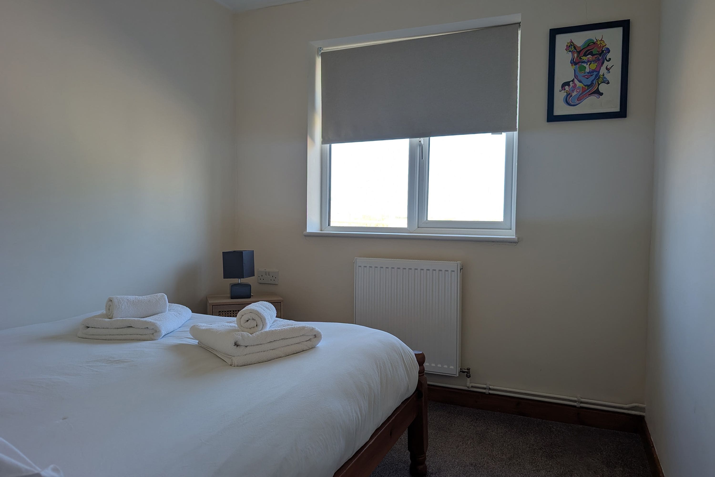 A bedroom with a bed, a window with a blind, a radiator, a nightstand with a lamp, and a framed picture on the wall.