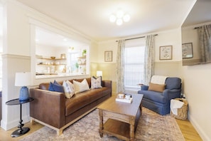 Een woonkamer met een bruine leren bank, een blauwe fauteuil, een houten salontafel en een bijzettafeltje met een lamp.